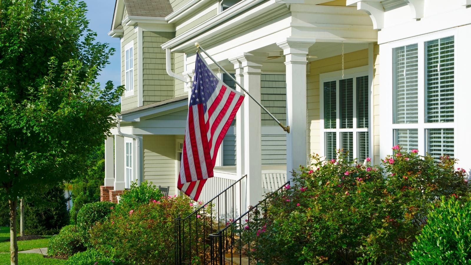 Tan house with an American flag hanging off the porch. 